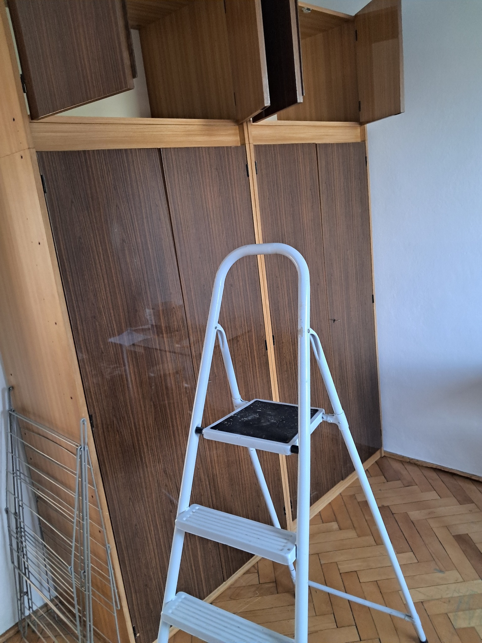 Wysoka szafa z otwartymi drzwiami, drabina malarska i suszarka na pranie w pokoju z parkietem. Przygotowania do prac wykończeniowych.