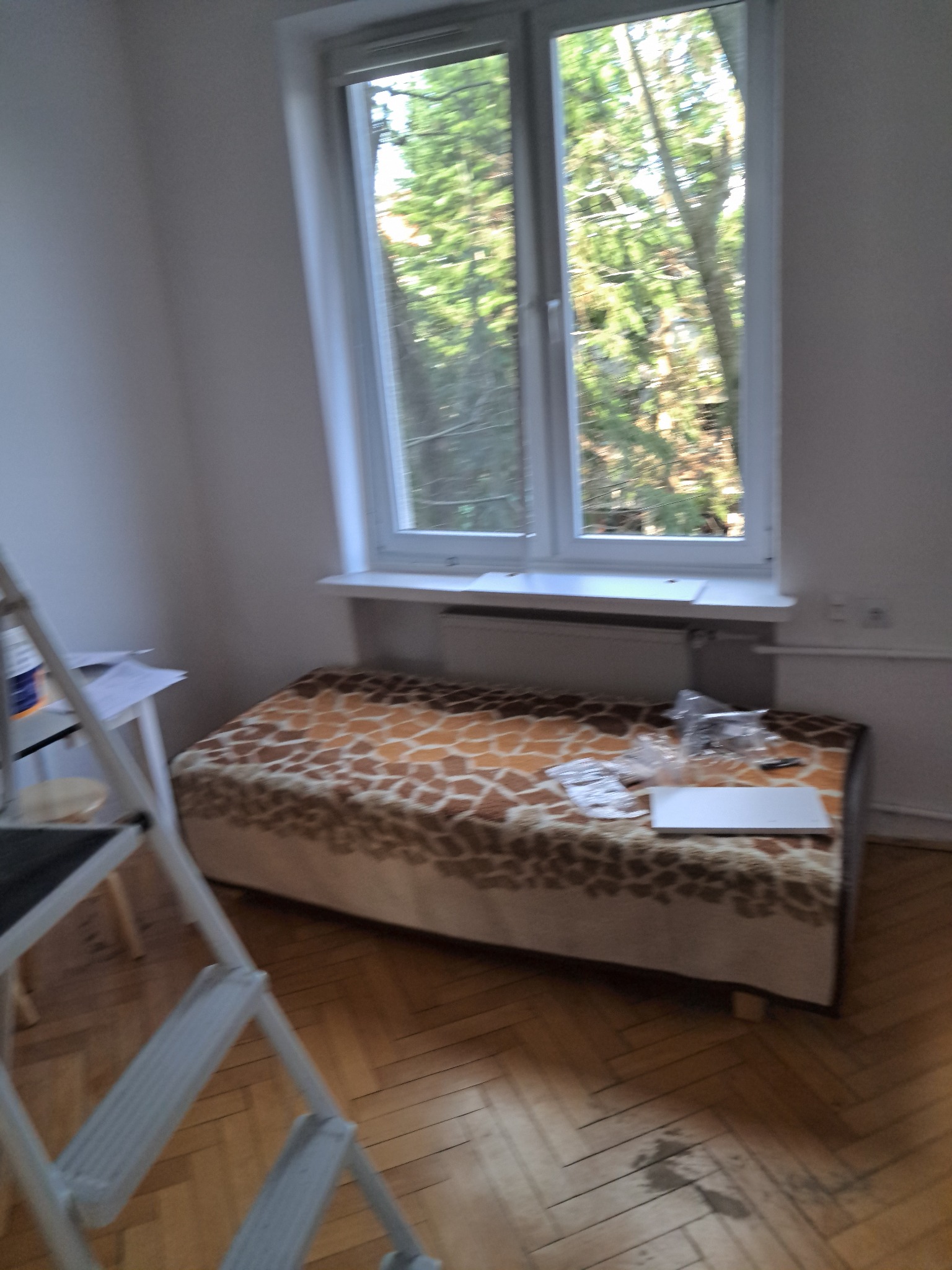 Pokój w trakcie remontu: drabina malarska, podłoga parkiet, okno z widokiem na zieleń, na podłodze rozłożone elementy mebli do montażu. Przygotowanie do prac wykończeniowych.