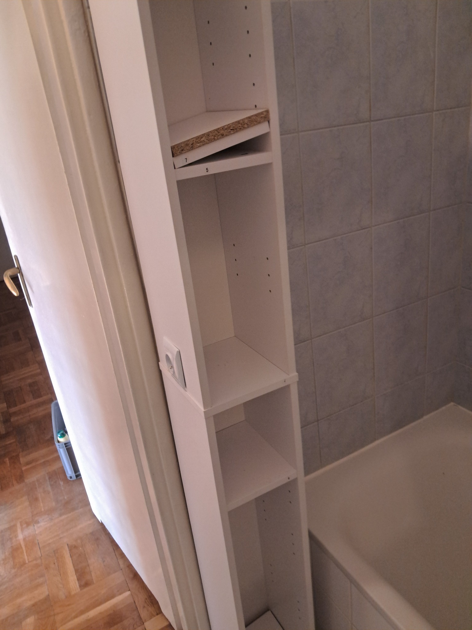 Wąska, biała szafka łazienkowa z półkami, częściowo zamontowana, widoczna obok wanny i ściany z szarych płytek. Ujęcie z bliska, z widocznym gniazdkiem elektrycznym.