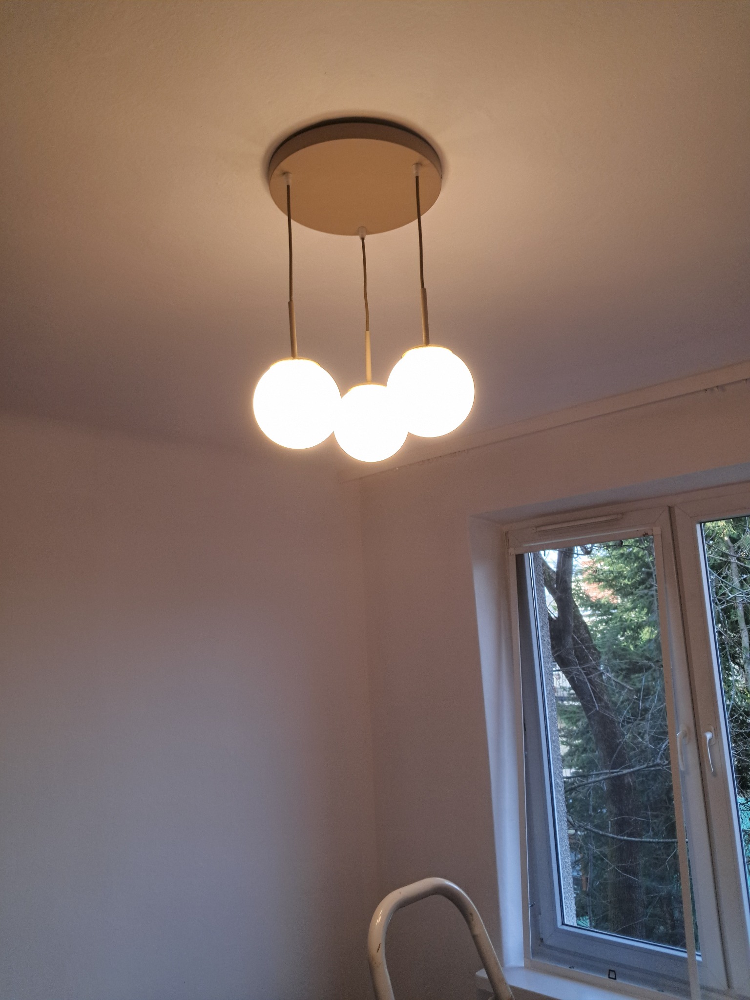 Zamontowana lampa sufitowa z trzema kulistymi kloszami w pokoju z oknem i fragmentem drabiny malarskiej. Minimalistyczny design oświetlenia.