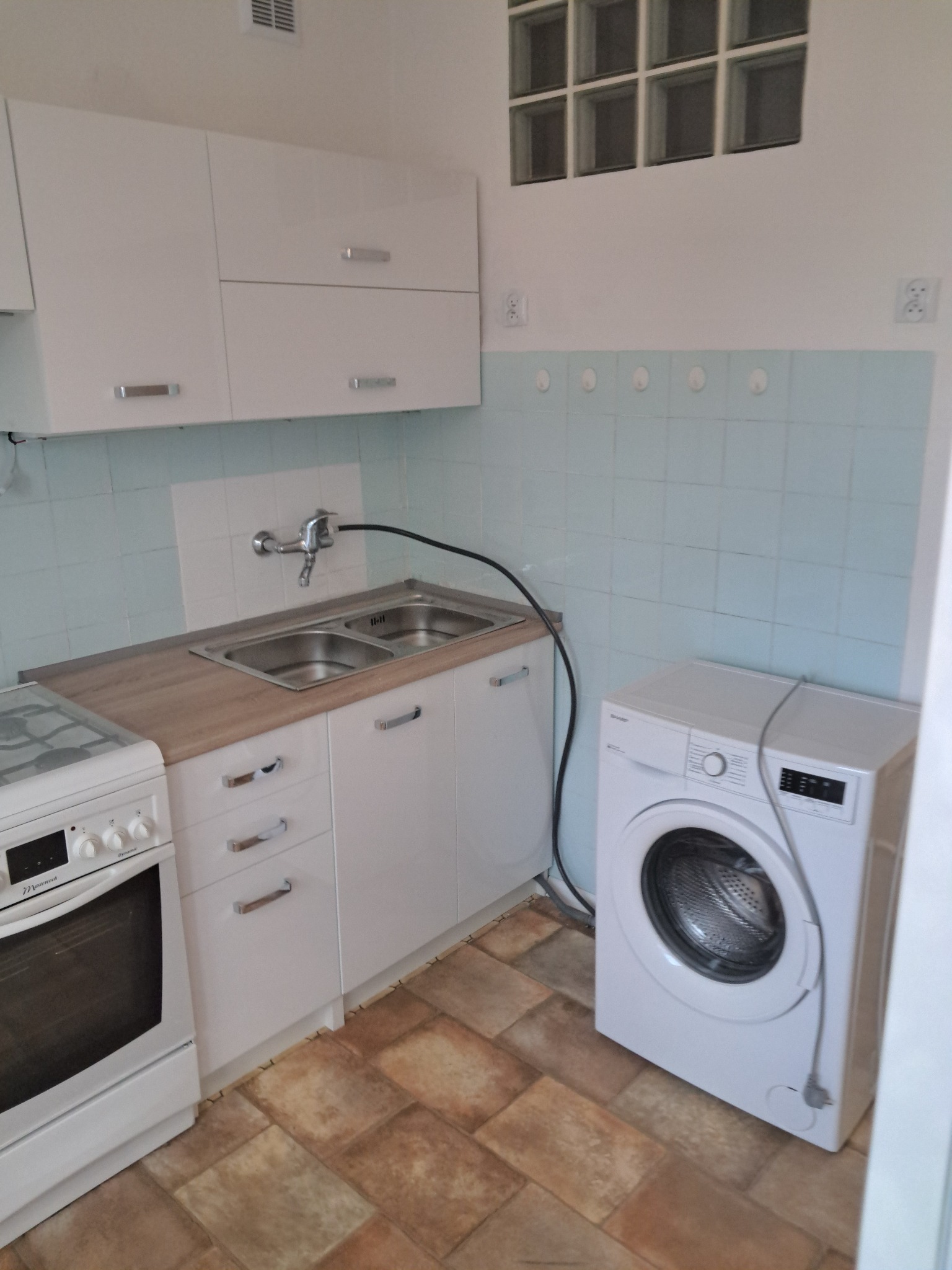 Jasna kuchnia po remoncie: białe szafki, beżowy blat, płytki w odcieniach błękitu, pralka i kuchenka. Widok na odnowione wnętrze z perspektywy podłogi.