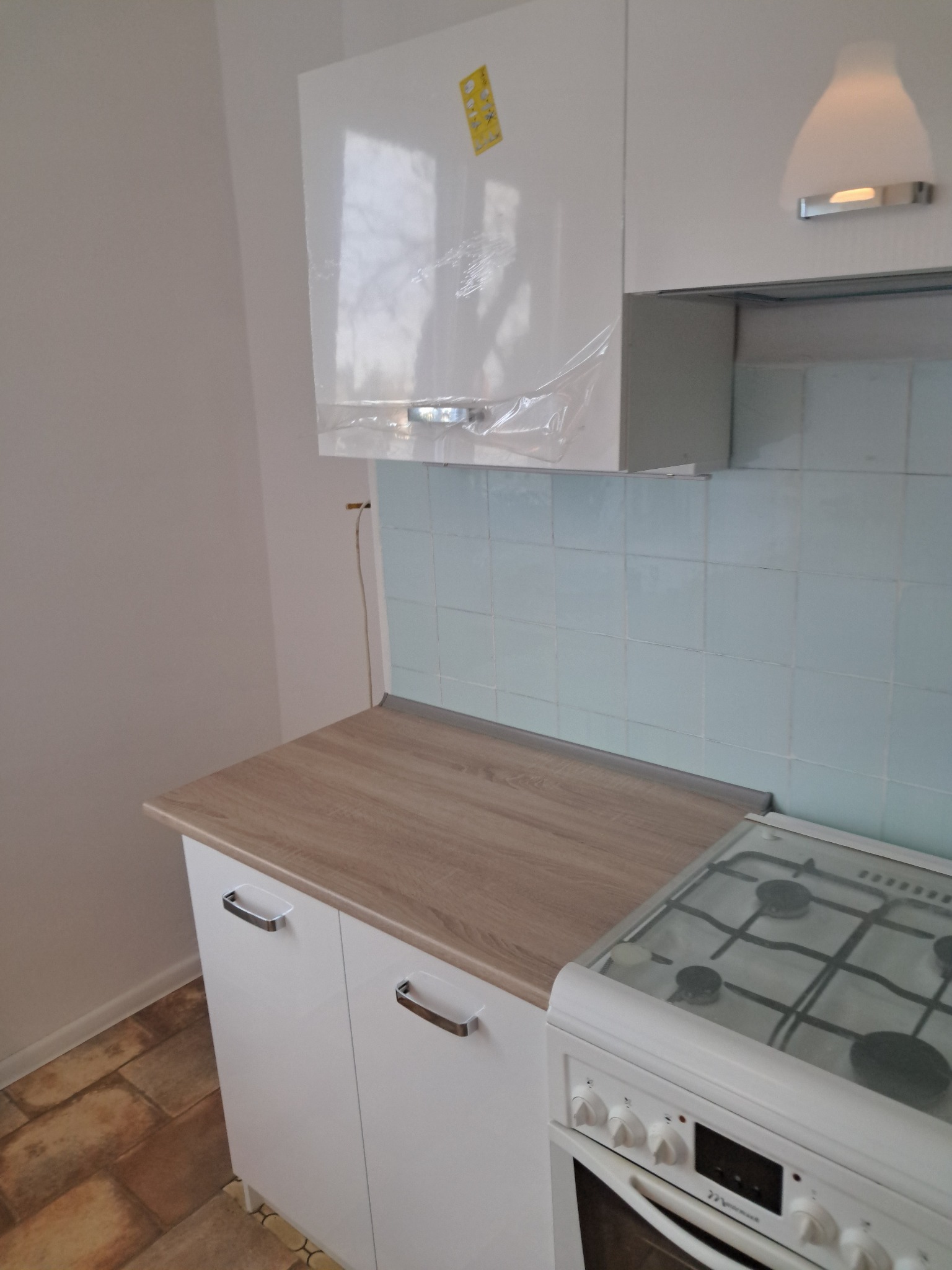 Jasna kuchnia z białymi szafkami i drewnianym blatem. Uwagę zwraca kuchenka gazowa z przezroczystą pokrywą i niebieskie płytki na ścianie.