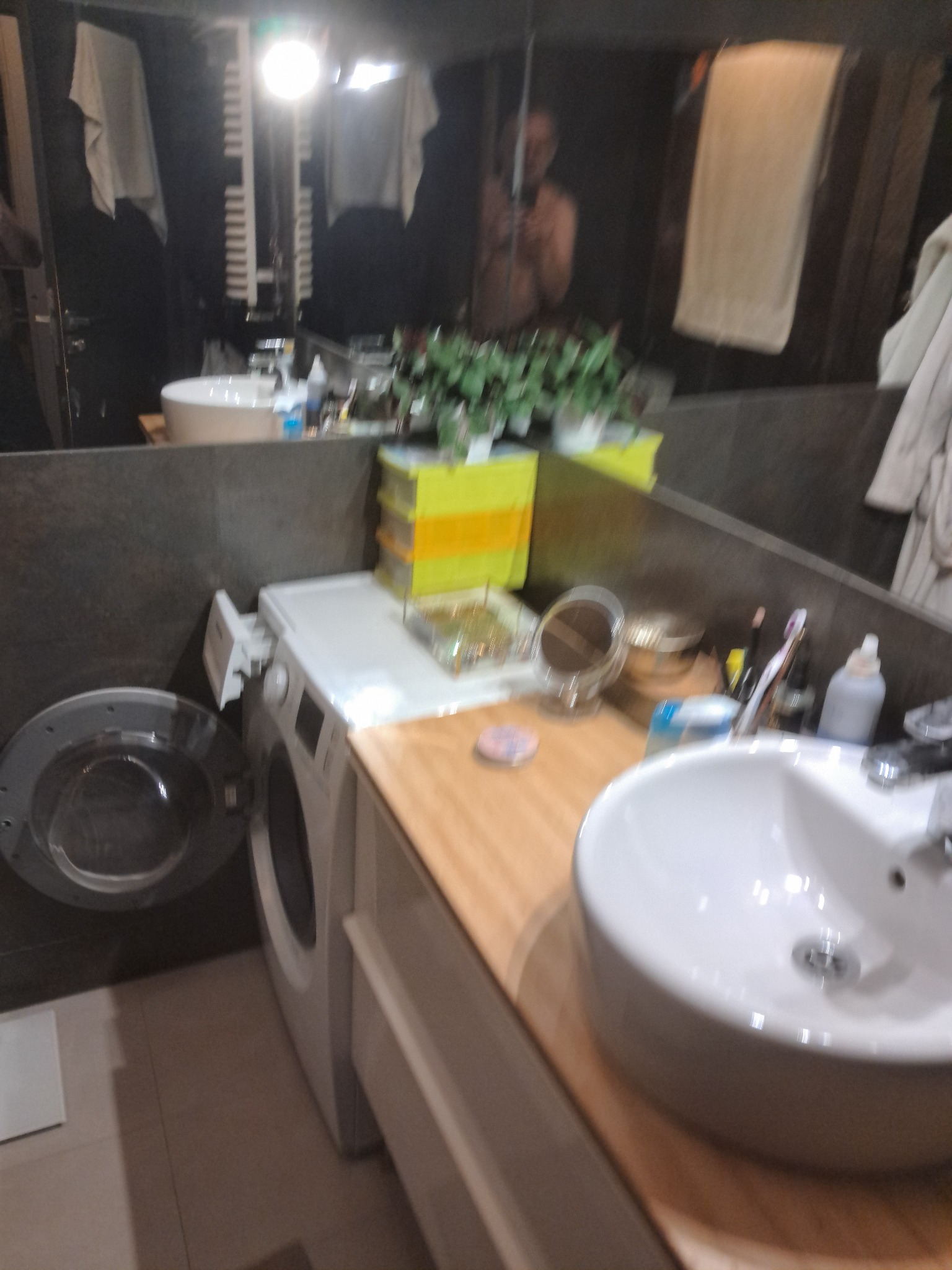 Nowoczesna łazienka z umywalką nablatową, pralką i dużym lustrem, w którym odbija się osoba. Ciemne płytki i drewniany blat dodają elegancji.