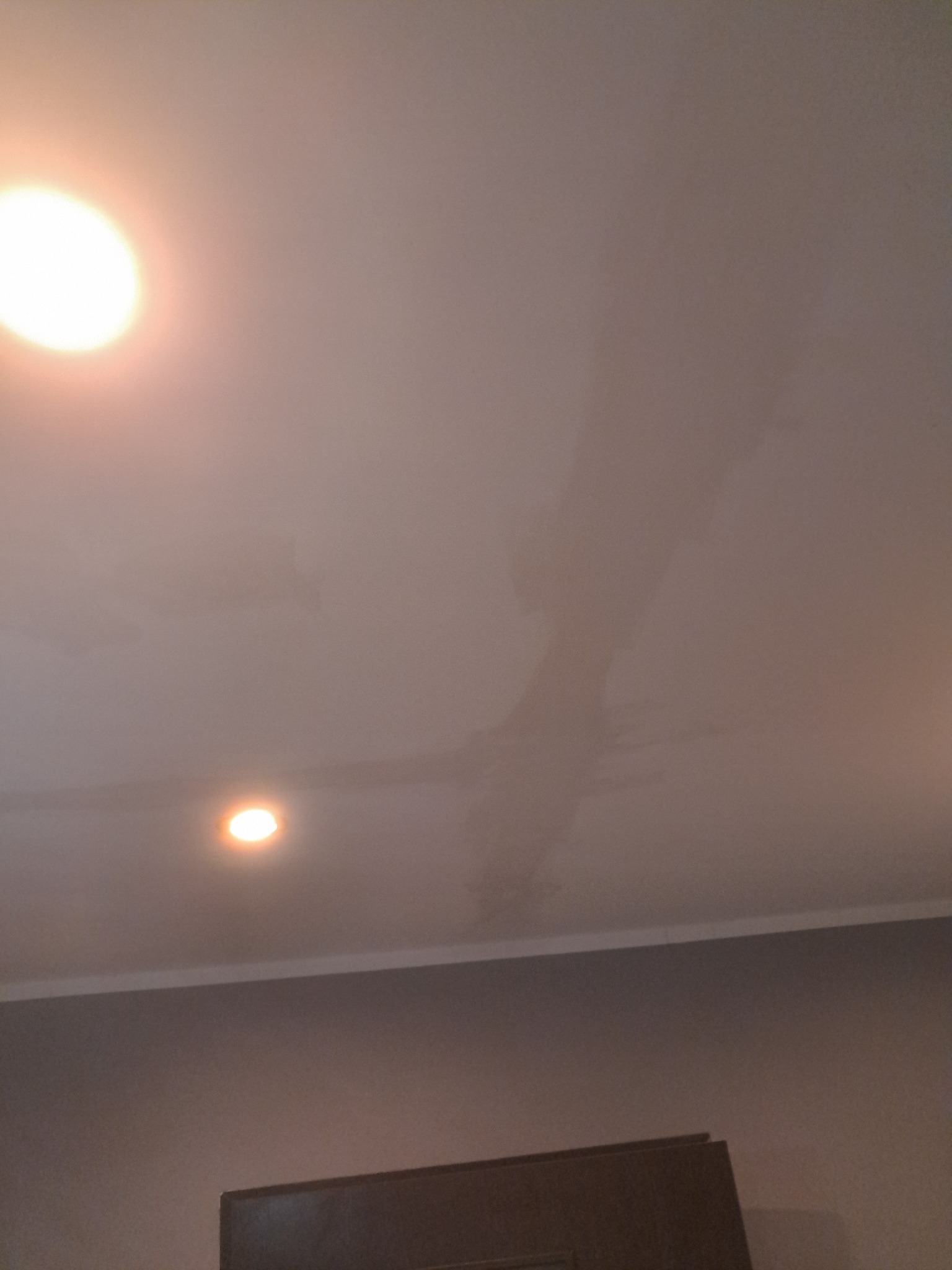 Biały sufit z widocznymi plamami i dwoma punktowymi źródłami światła, fragment szarej ściany i mebla w dolnej części kadru. Niejednolita powierzchnia sufitu.