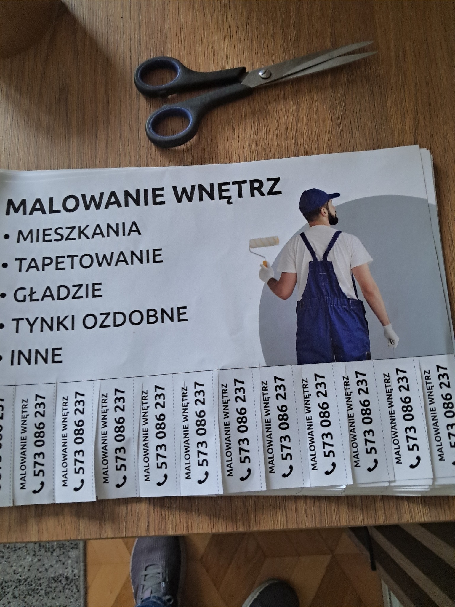 Ulotka reklamowa z hasłem 'Malowanie Wnętrz', wykazem usług i numerem telefonu, leżąca na stole obok nożyczek. Widoczne fragmenty butów osoby fotografującej.