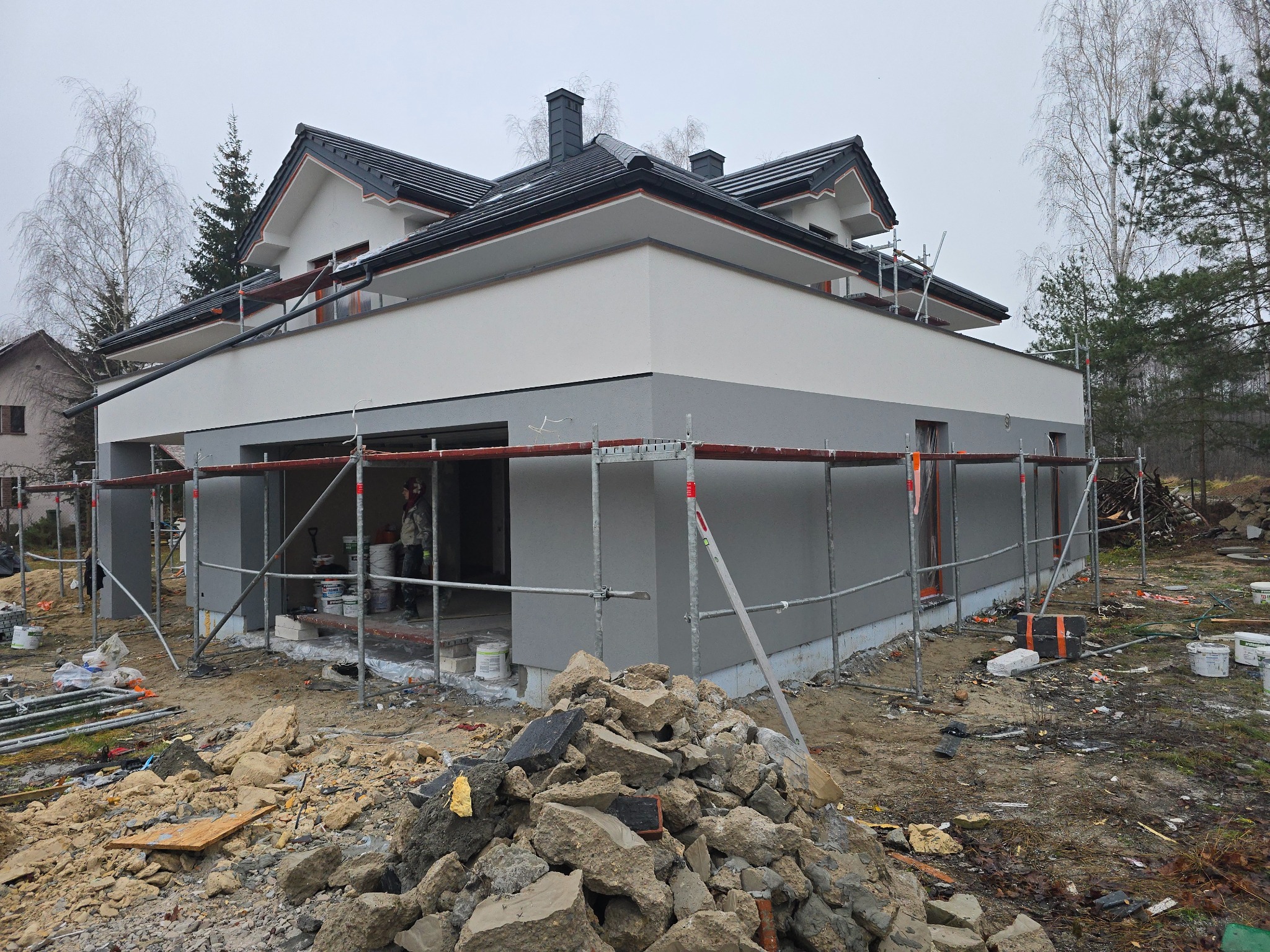Dwukondygnacyjny dom w trakcie tynkowania elewacji, z widocznymi rusztowaniami i stertą gruzu na pierwszym planie. Częściowo otynkowana fasada w odcieniach szarości i bieli.