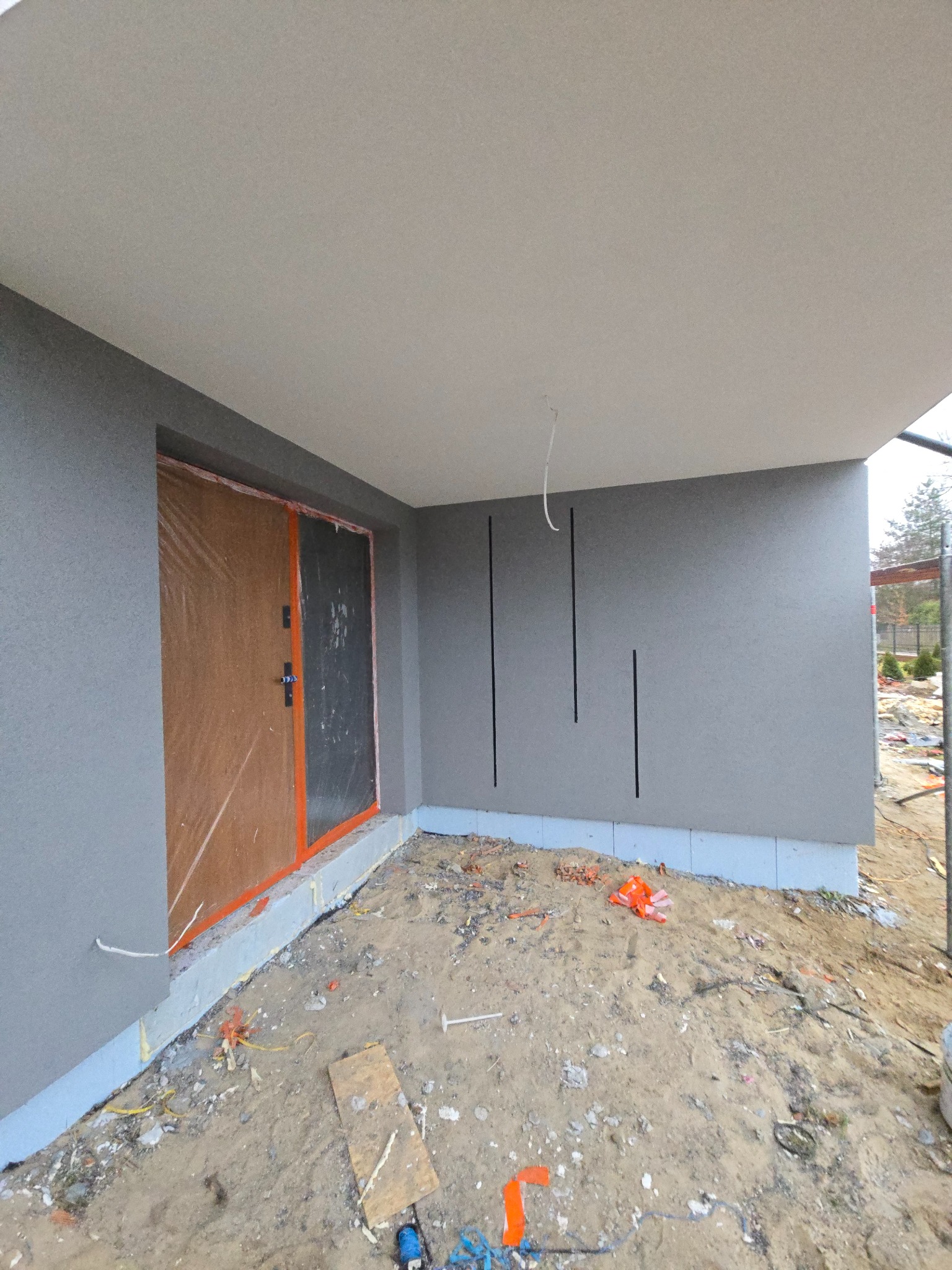 Szara elewacja budynku w trakcie wykańczania, z widocznymi przewodami i zabezpieczonymi drzwiami wejściowymi folią ochronną. Teren wokół w nieładzie.