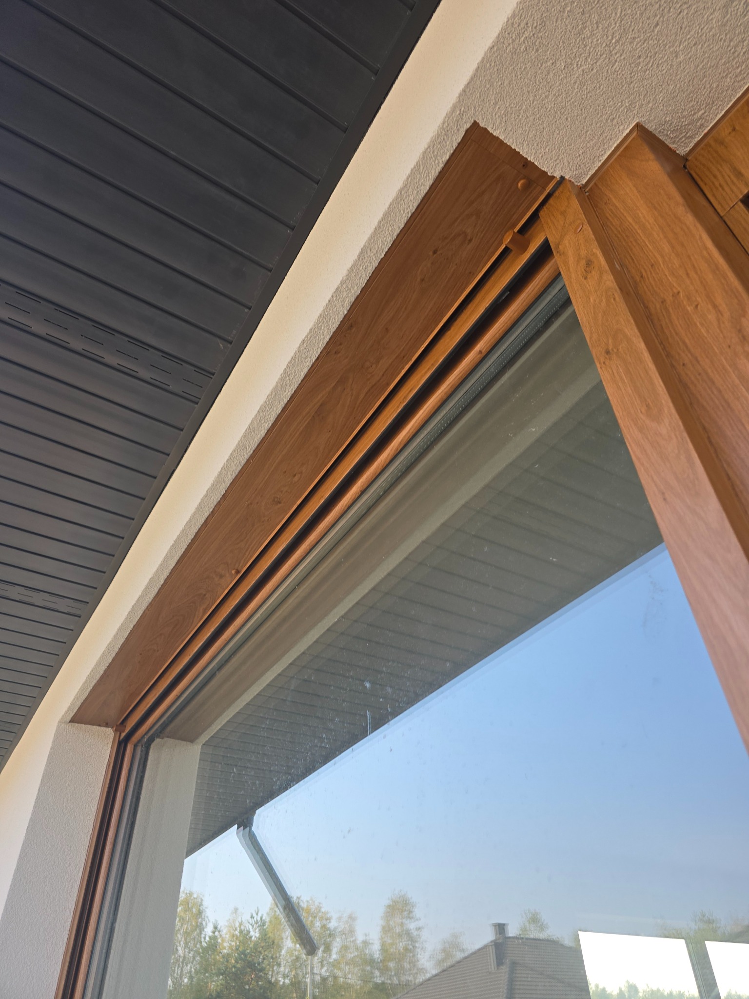 Fragment elewacji z oknem o drewnopodobnej ramie, widoczny fragment ocieplenia i podbitki dachowej. Nowoczesne wykończenie.