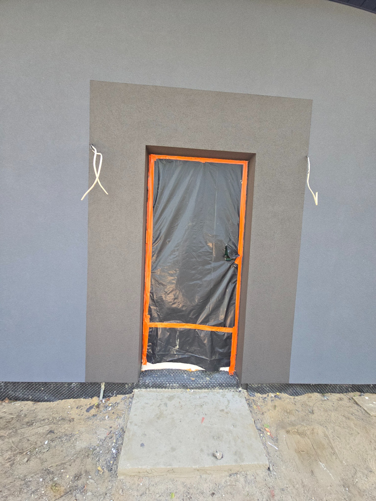 Elewacja budynku w Żyrardowie w trakcie prac. Widoczny fragment otynkowanej ściany z zabezpieczonym wejściem folią i taśmą. Dwa odcienie szarości na fasadzie.