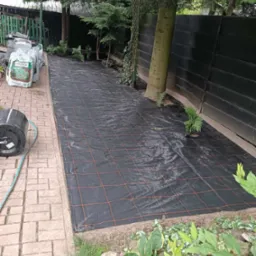 Rozłożona czarna agrotkanina z pomarańczową siatką na przygotowanym podłożu w ogrodzie, obok worki z korą i wąż do podlewania. Widoczne drzewa i drewniany płot.