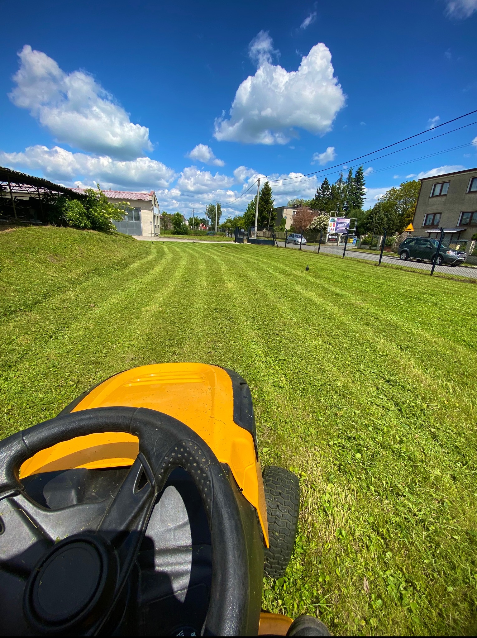 Żółty traktorek do koszenia trawy w trakcie pracy, widok z perspektywy operatora na świeżo skoszoną trawę w pasy, błękitne niebo z chmurami w tle.