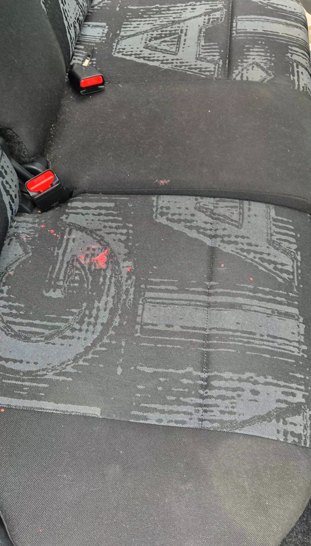 Zabrudzone krwią ciemne, materiałowe siedzenie samochodowe z widocznymi plamami i zapięciem pasów. Wzór geometryczny na tapicerce.