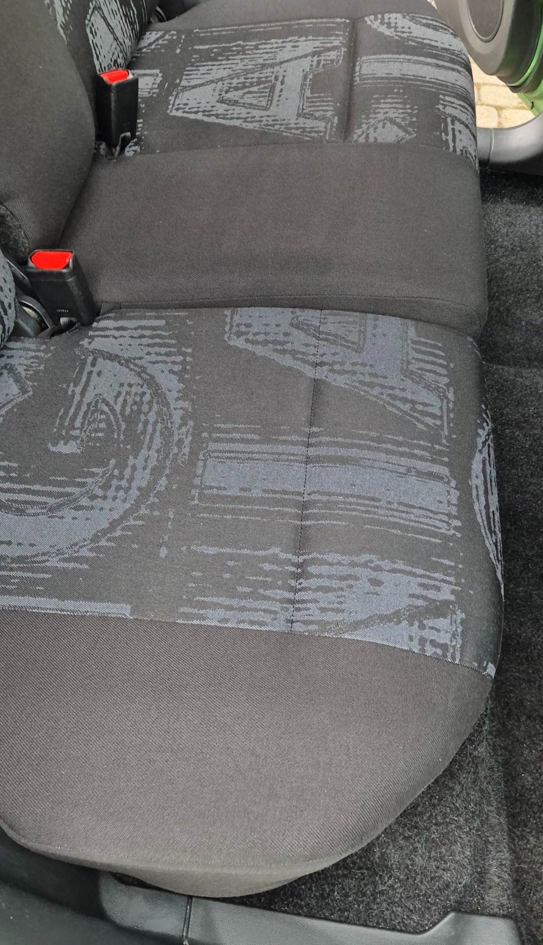 Szczegółowe ujęcie tylnej kanapy samochodu z wzorem w odcieniach szarości i czerwonymi elementami pasów bezpieczeństwa. Widoczne ślady użytkowania na tapicerce.