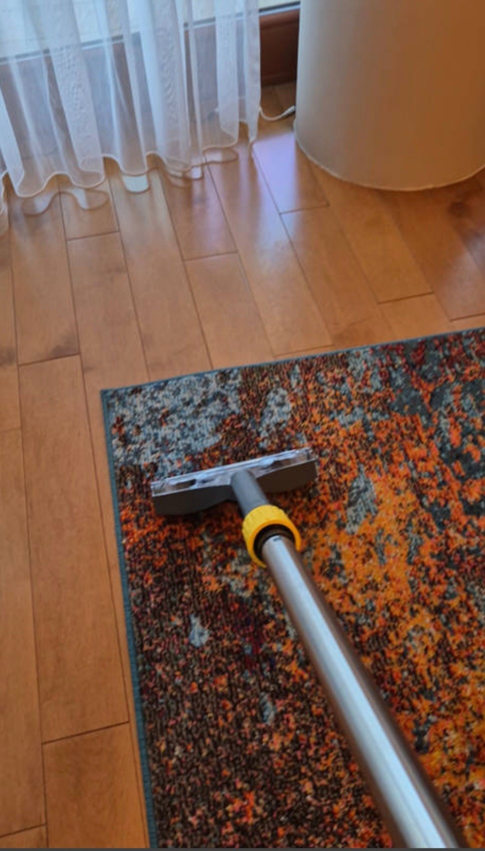 Czyszczenie wzorzystego dywanu na podłodze z desek za pomocą odkurzacza piorącego z żółtymi elementami. Widoczne zasłony i lampa w tle.