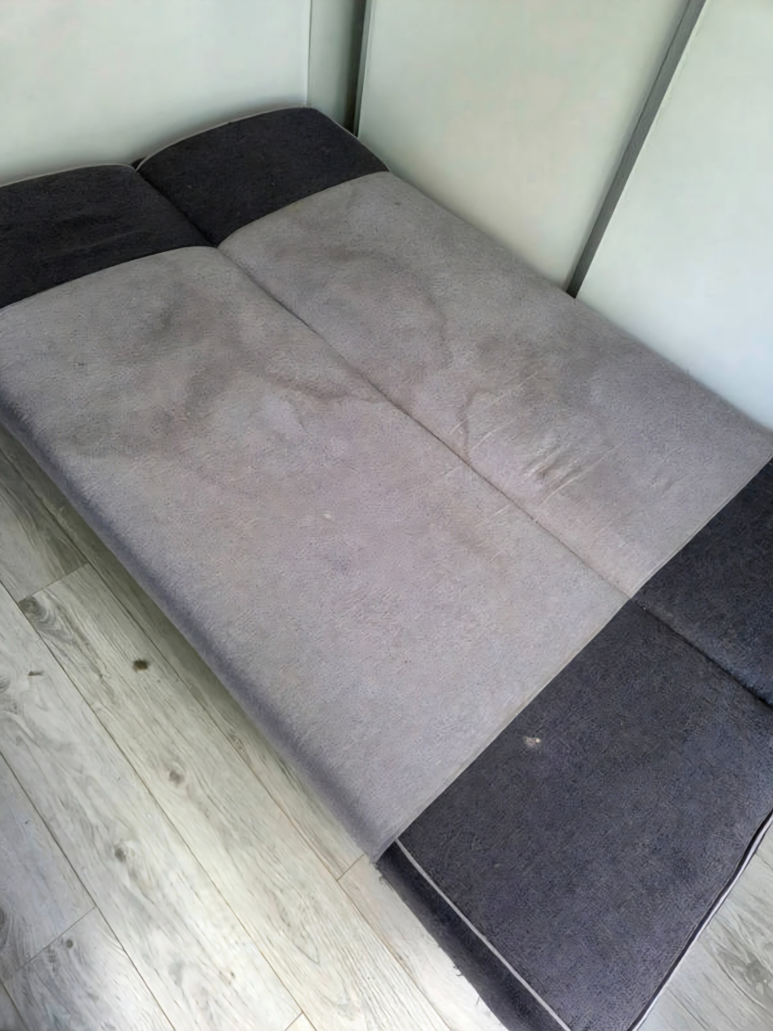 Szaro-grafitowa rozkładana kanapa z widocznymi plamami na siedzisku, ustawiona w rogu pokoju na jasnej podłodze. Widoczne zabrudzenia na tapicerce.