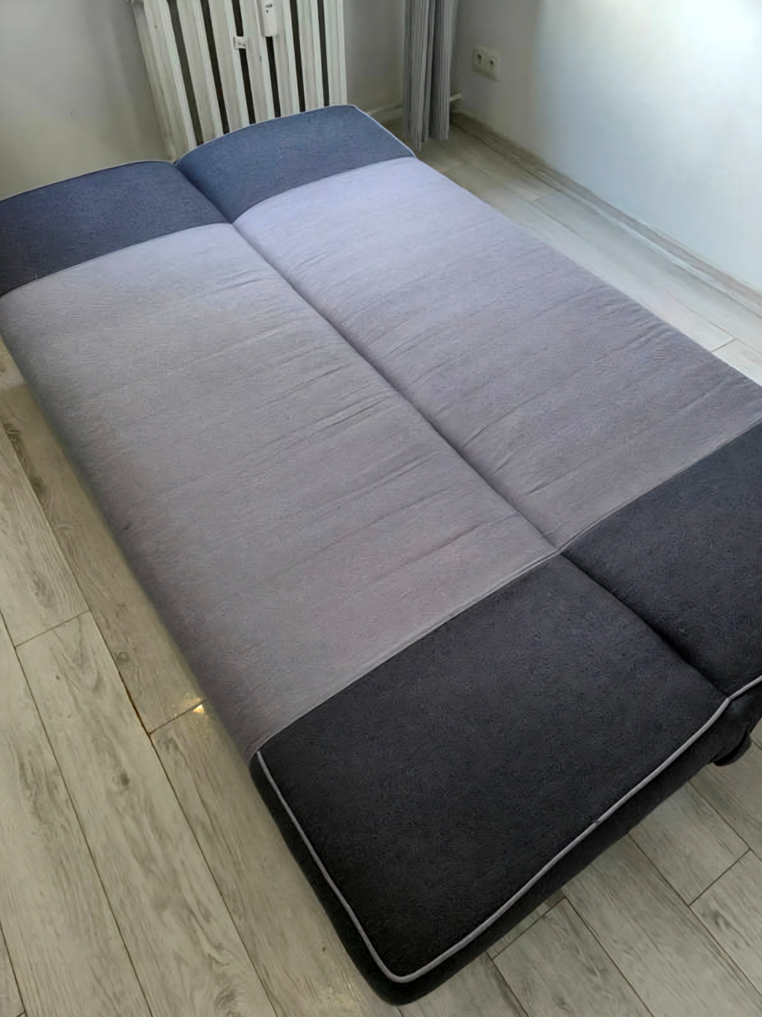 Rozłożona, dwukolorowa kanapa z szarym siedziskiem i grafitowymi bokami, ustawiona na drewnianej podłodze w jasnym pomieszczeniu. Widoczny grzejnik i zasłona w tle.