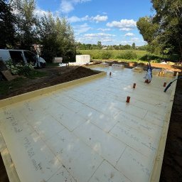 LITBUD - Ocieplone fundamenty pod budowę domu szkieletowego z widocznymi elementami instalacji, na tle zieleni i błękitnego nieba z chmurami.