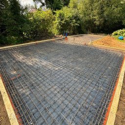 LITBUD - Zbrojenie płyty fundamentowej na czarnej folii, obramowane szalunkiem z desek. W tle zieleń drzew i krzewów. Widok z góry, szeroki kadr.