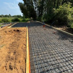 LITBUD - Zbrojenie fundamentu pod dom na działce z zielenią w tle. Widoczne szalunki i wystające rury instalacyjne. Prace budowlane w toku, przygotowanie do wylewki.