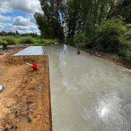 LITBUD - Świeżo wylana, gładka płyta betonowa pod budowę domu na działce z zielenią w tle i pracownikiem w oddali. Widoczne wystające pręty zbrojeniowe.