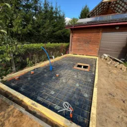 Zbrojenie płyty fundamentowej z instalacjami w Hajnówce. Siatka zbrojeniowa na czarnej folii, drewniany szalunek, widoczny fragment budynku z panelami słonecznymi.