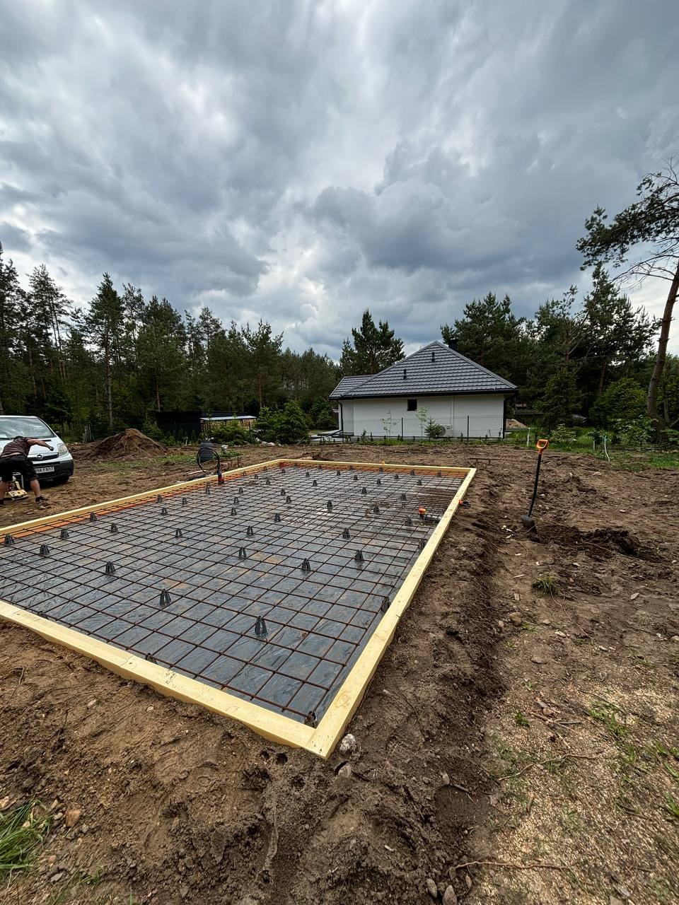 Zbrojenie płyty fundamentowej pod budynek, zabezpieczone folią, obramowane drewnianą konstrukcją. W tle dom jednorodzinny i pochmurne niebo.