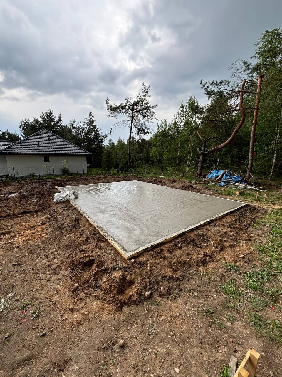 Wylany fundament pod budowę domu, otoczony drewnianym szalunkiem. W tle widoczny dom jednorodzinny i zieleń drzew. Pochmurne niebo.