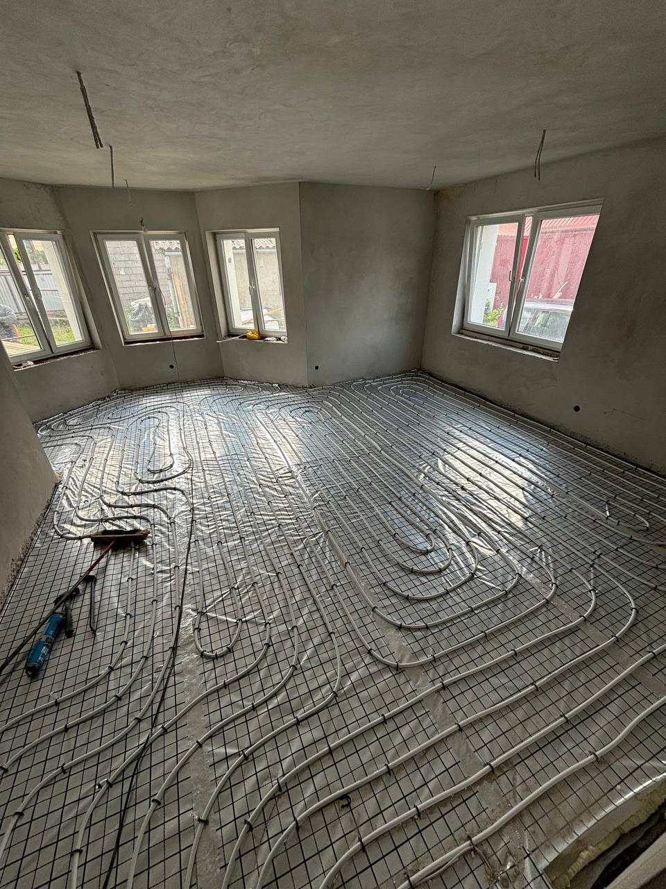 Białe rurki ogrzewania podłogowego ułożone na czarnej siatce w surowym pomieszczeniu z trzema oknami. Widoczne narzędzia pracy.