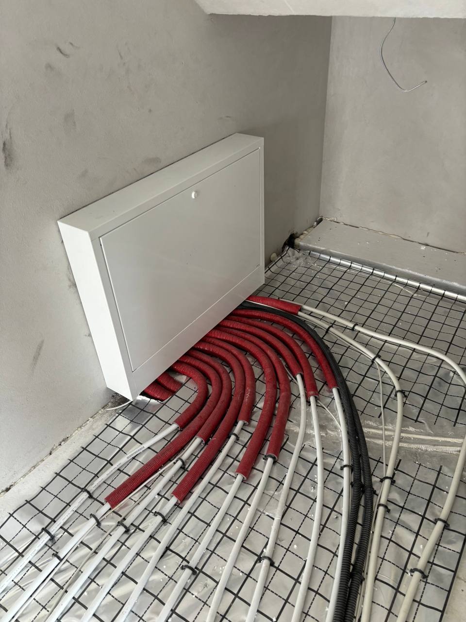 Rozłożone rury ogrzewania podłogowego, częściowo w czerwonej osłonie, wychodzące z białej skrzynki na podłodze pokrytej siatką, surowy beton na ścianach.