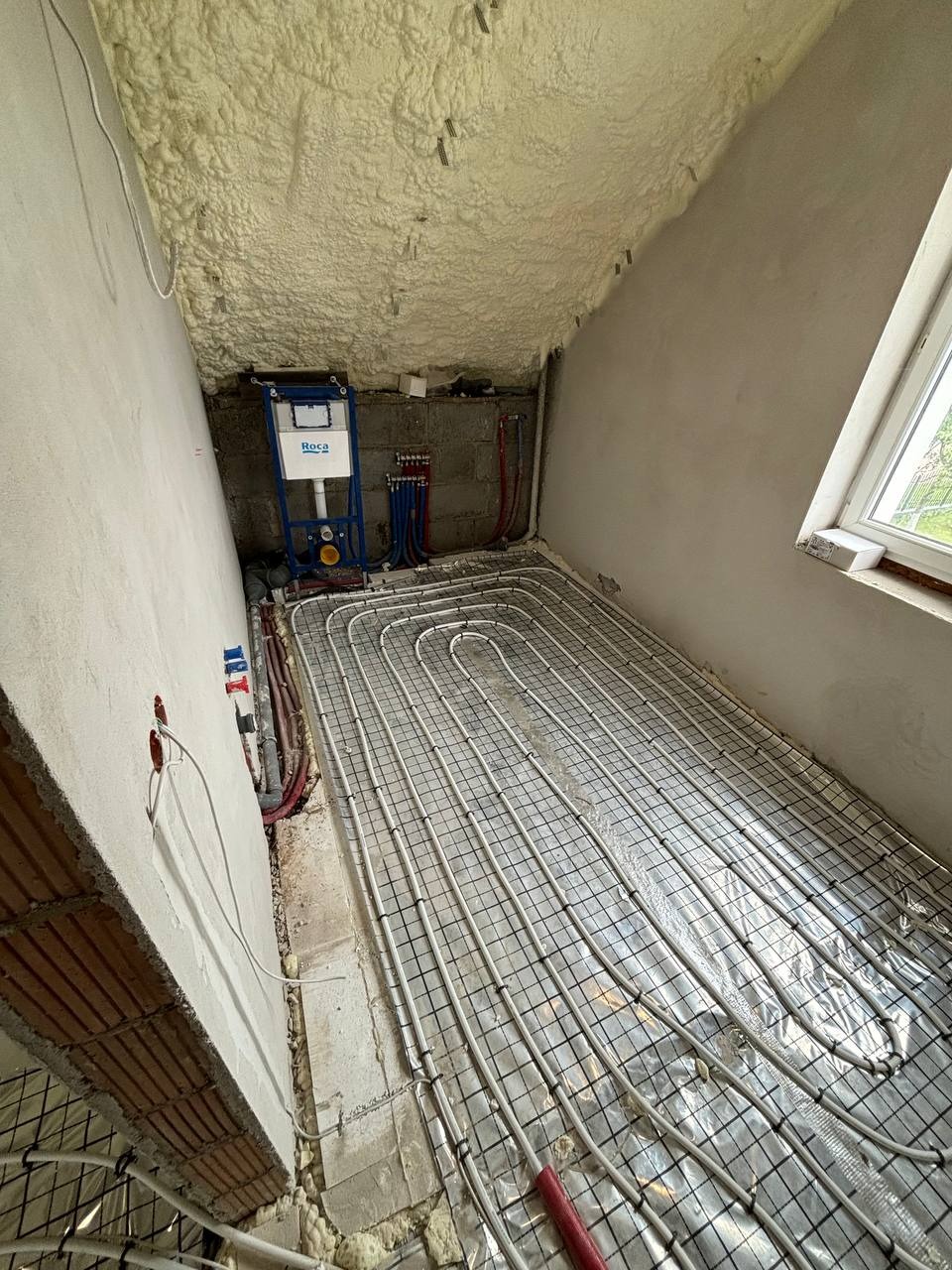 Instalacja ogrzewania podłogowego na folii aluminiowej w pokoju na poddaszu. Widoczny rozdzielacz i pianka izolacyjna na suficie.