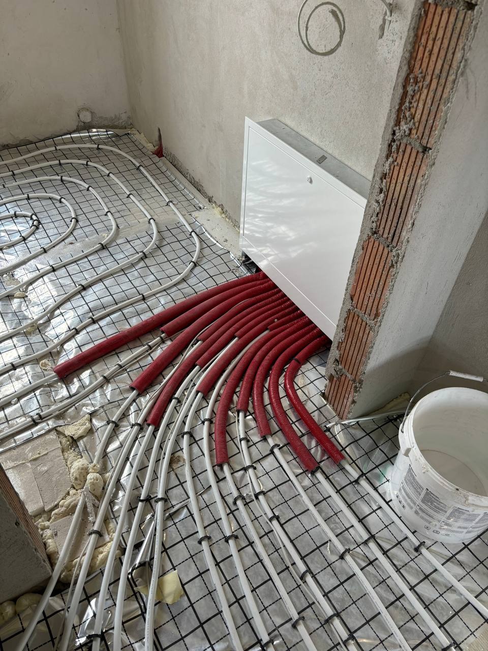 Instalacja ogrzewania podłogowego z czerwonymi rurami w folii aluminiowej, widok z góry na surową posadzkę, biała skrzynka rozdzielcza przy ścianie i wiadro farby.