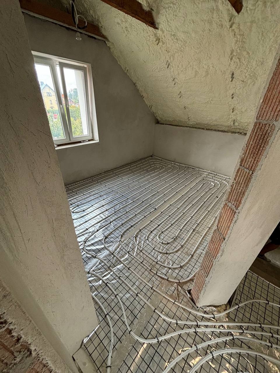 Instalacja ogrzewania podłogowego na poddaszu z oknem, widoczne rury na folii i siatce, ściany ocieplone pianą, surowy stan wykończenia.