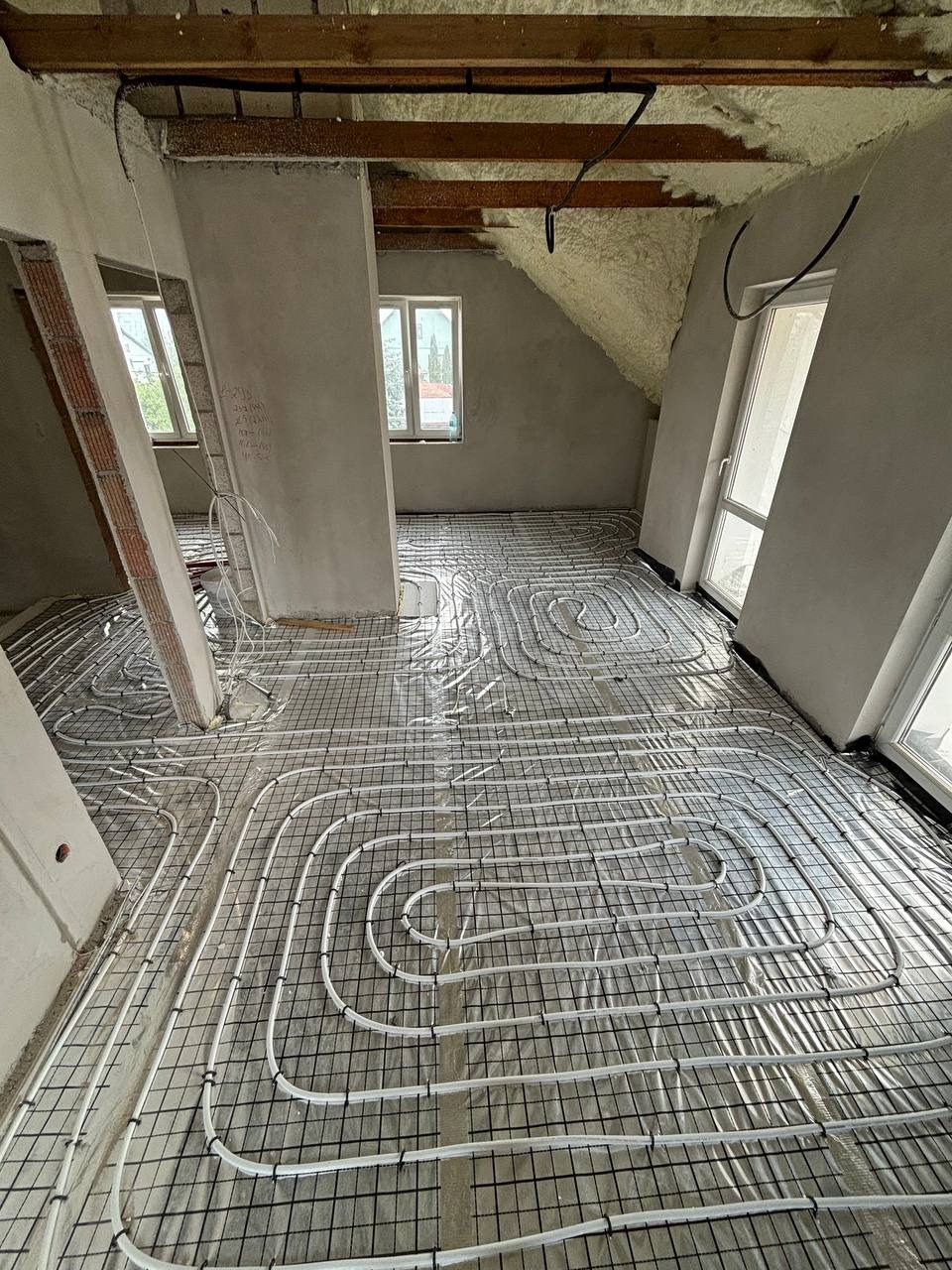 Białe rury ogrzewania podłogowego ułożone na czarnej siatce, w surowym wnętrzu poddasza z oknami i pianą izolacyjną na skosach dachu.