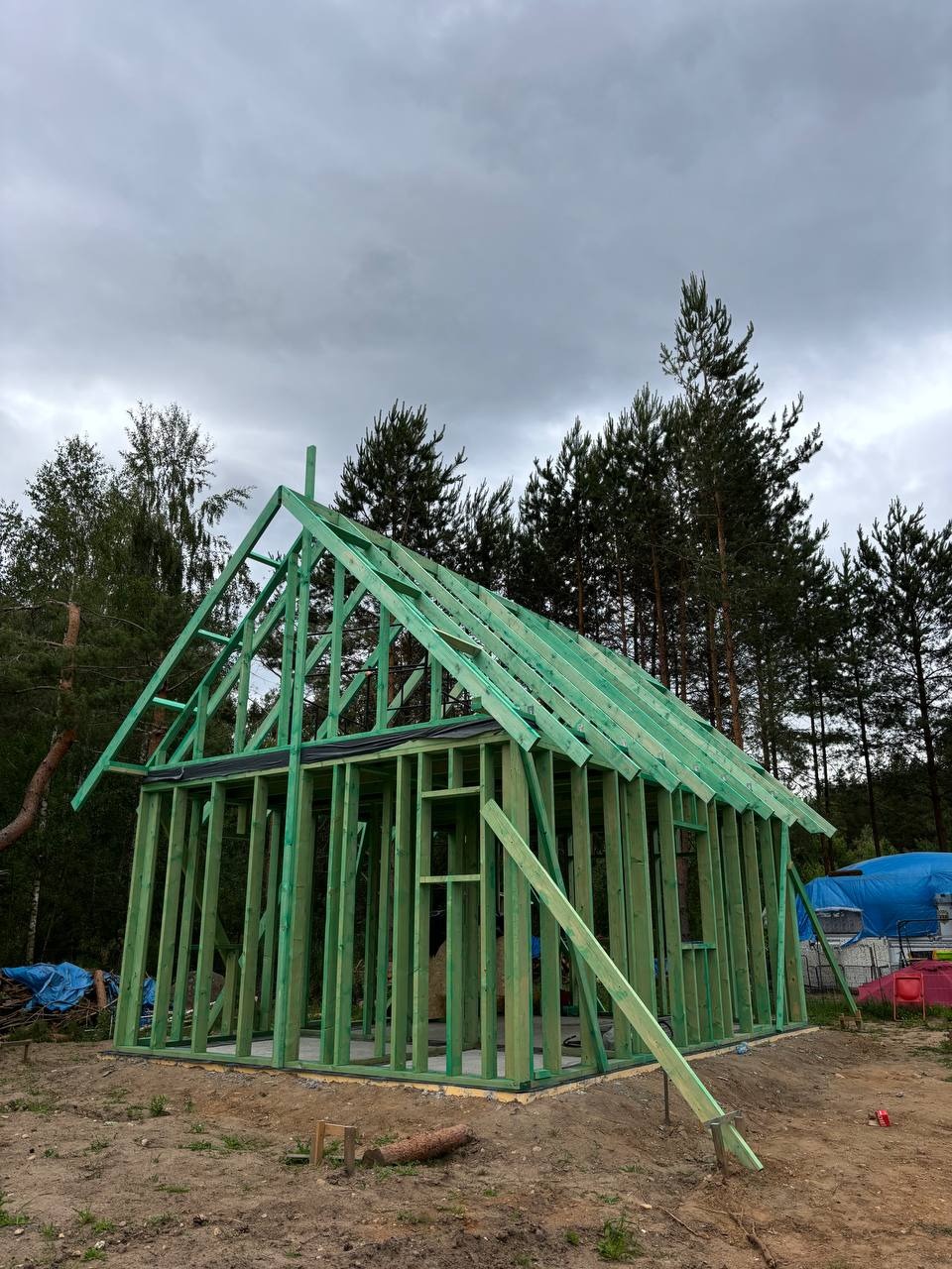 Surowy szkielet domu w lesie, zielone drewno konstrukcyjne, pochmurne niebo, widoczne drzewa w tle. Budowa w toku, ziemia wokół.