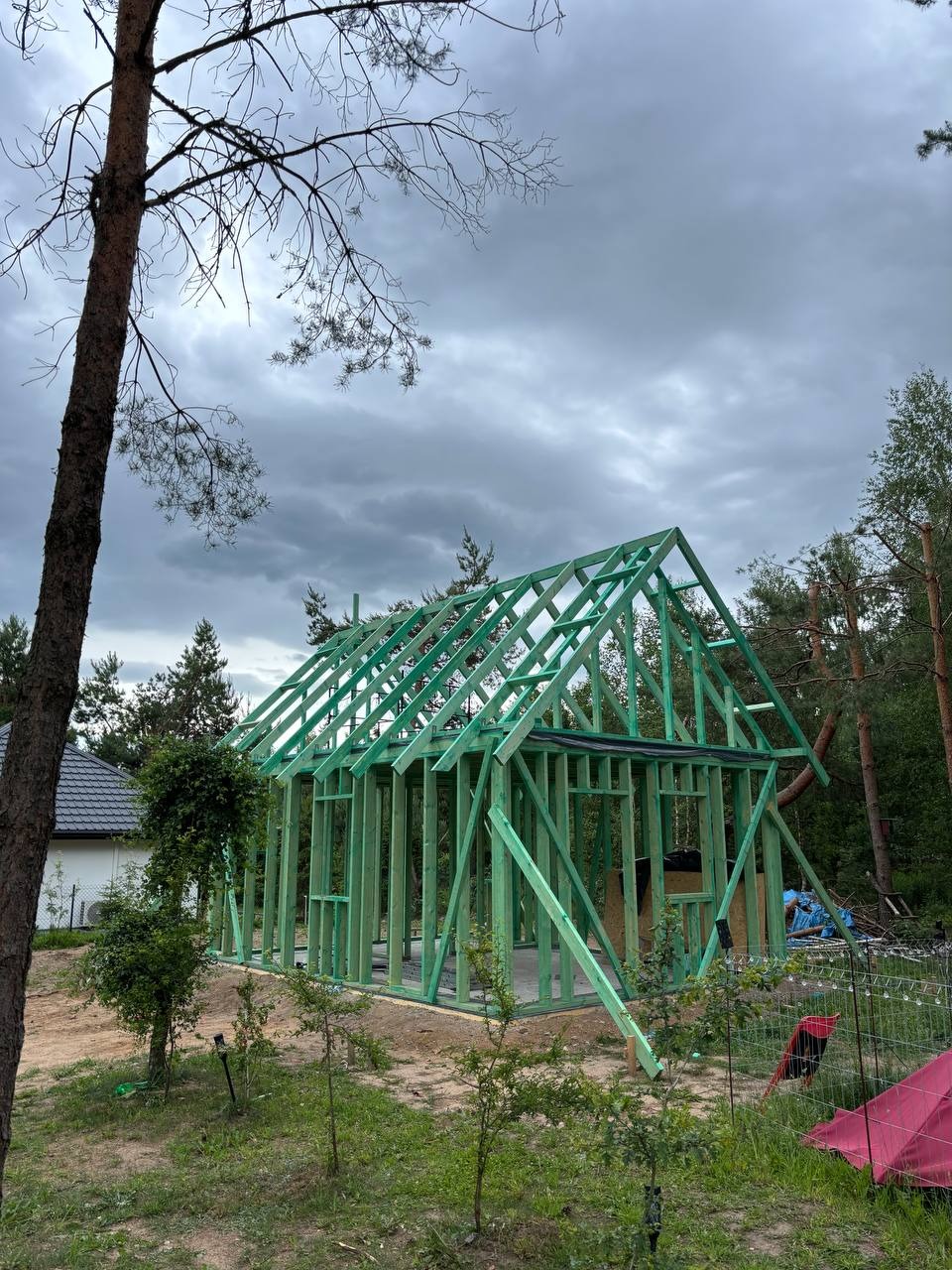 Surowy szkielet domu z zielonego drewna pod pochmurnym niebem, otoczony drzewami i zielenią. Widoczne detale konstrukcyjne i etap budowy.