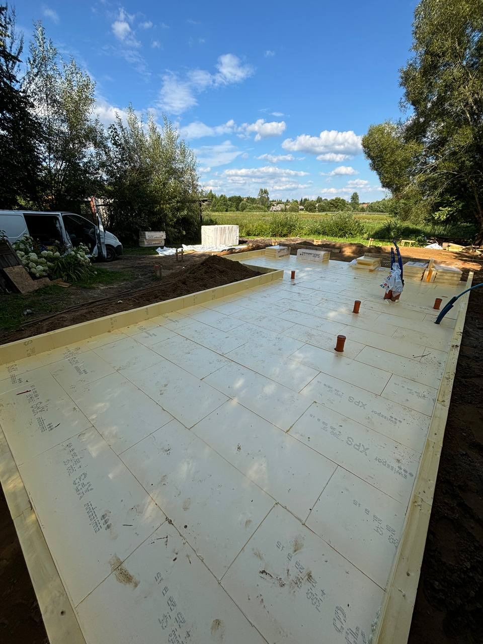 Ocieplone fundamenty pod budowę domu szkieletowego z widocznymi elementami instalacji, na tle zieleni i błękitnego nieba z chmurami.