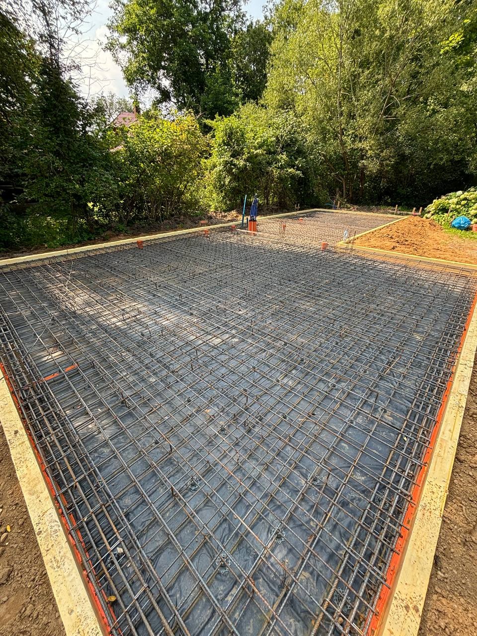 Zbrojenie płyty fundamentowej na czarnej folii, obramowane szalunkiem z desek. W tle zieleń drzew i krzewów. Widok z góry, szeroki kadr.
