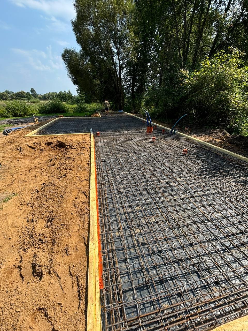 Zbrojenie fundamentu pod dom na działce z zielenią w tle. Widoczne szalunki i wystające rury instalacyjne. Prace budowlane w toku, przygotowanie do wylewki.