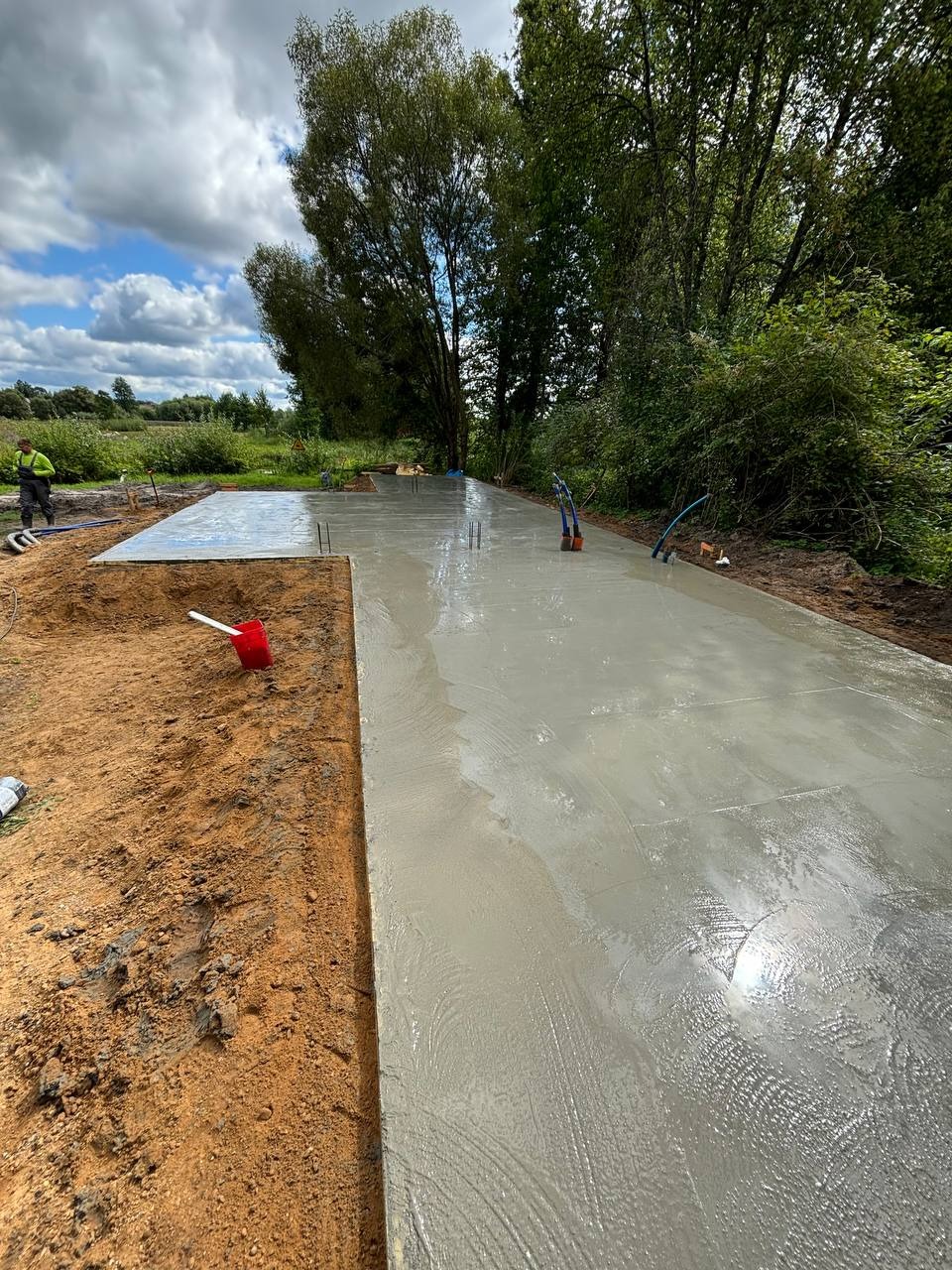 Świeżo wylana, gładka płyta betonowa pod budowę domu na działce z zielenią w tle i pracownikiem w oddali. Widoczne wystające pręty zbrojeniowe.
