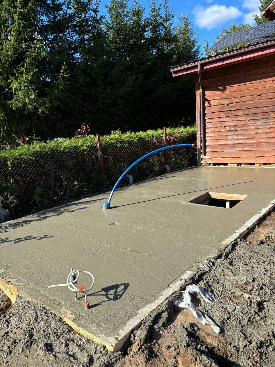 Świeżo wylana, szara płyta fundamentowa z widocznym otworem i niebieską rurą, otoczona ziemią. W tle drewniany budynek i zieleń.