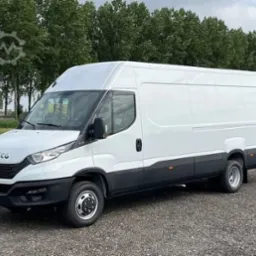 Biały, dostawczy Iveco Daily na żwirowym podłożu, otoczony zielenią. Widok z boku, lekko od przodu. Idealny do przeprowadzek i transportu.
