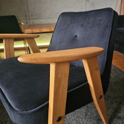 Dawid Dźwierzyński - Czarne, welurowe krzesło z drewnianymi podłokietnikami, widoczne z bliska. W tle szara ściana z dekoracyjnymi elementami i fragment lampy.