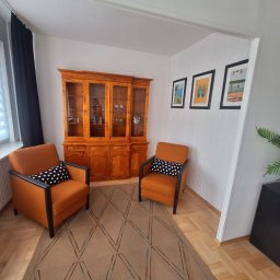 Dawid Dźwierzyński - Dwa pomarańczowe fotele z czarnymi podłokietnikami i poduszkami w kropki, ustawione naprzeciwko siebie na wzorzystym dywanie, w tle drewniany kredens i obrazy.