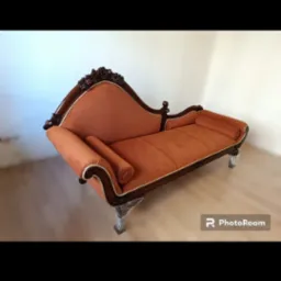 Stylowa leżanka tapicerowana pomarańczową tkaniną, z ciemnego drewna zdobieniami i eleganckimi nóżkami na tle jasnej ściany. Widoczny znak wodny PhotoRoom.
