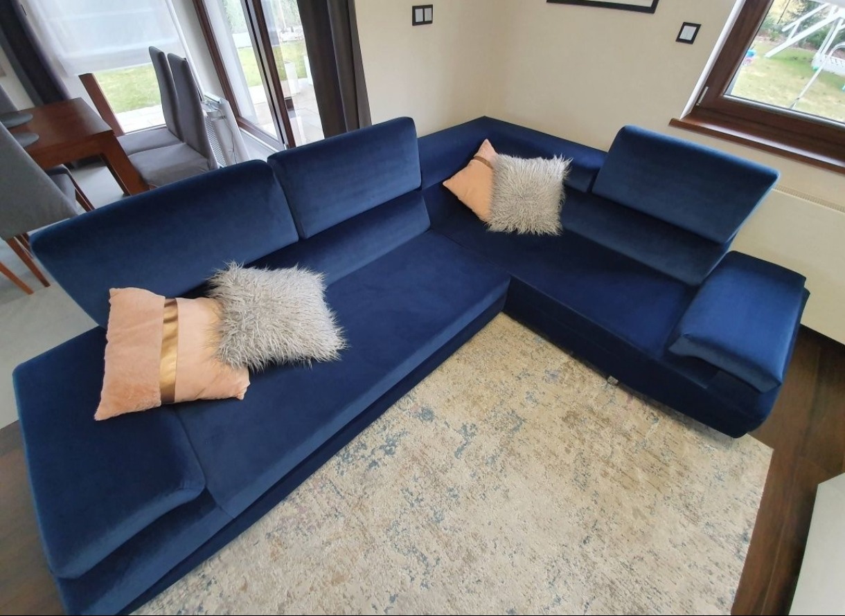 Narożnik tapicerowany, aksamitny w odcieniach granatu, z poduszkami w kolorach beżu i szarości, na wzorzystym dywanie. Ujęcie z góry, widok na salon z oknem.
