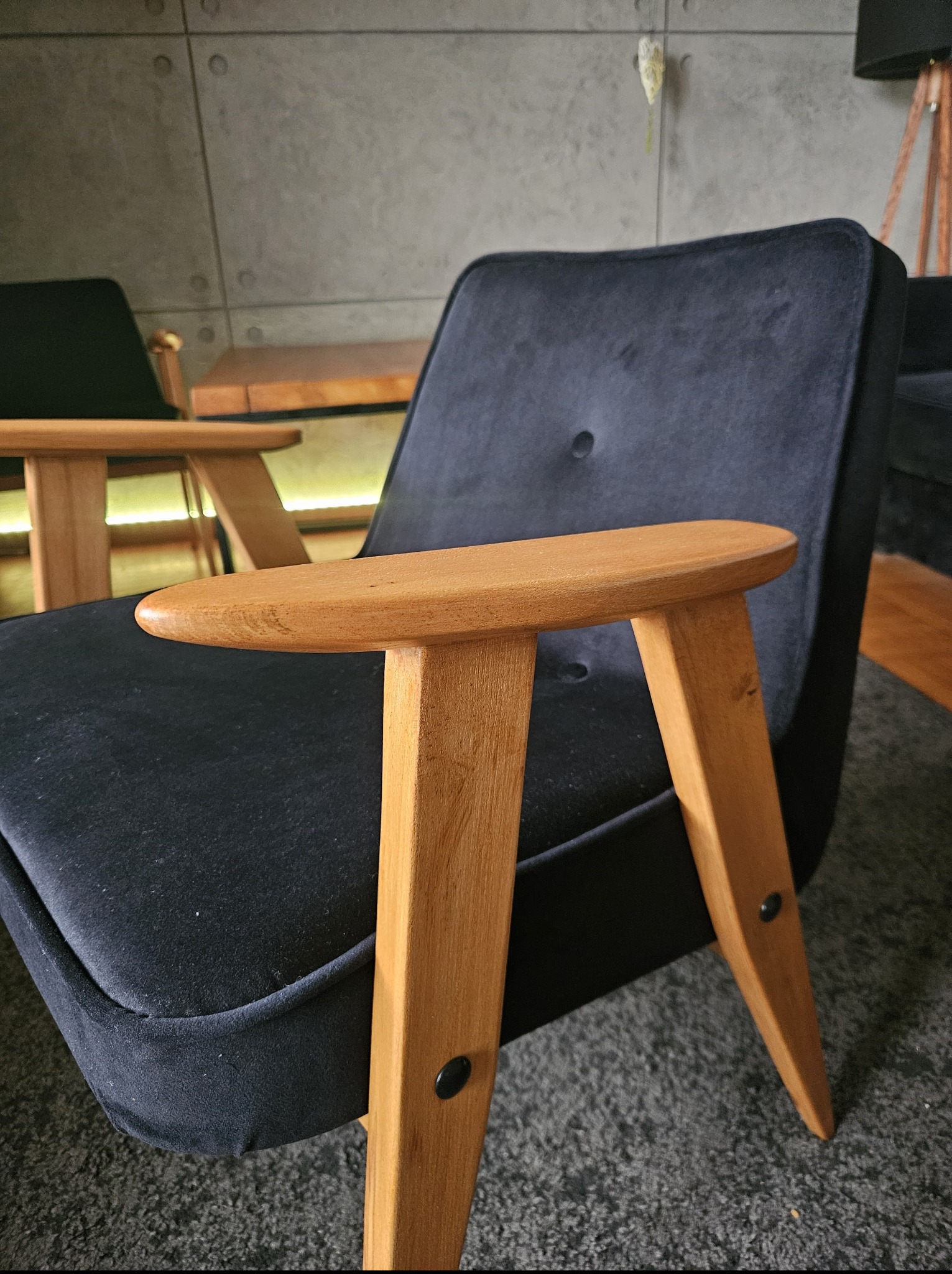 Czarne, welurowe krzesło z drewnianymi podłokietnikami, widoczne z bliska. W tle szara ściana z dekoracyjnymi elementami i fragment lampy.