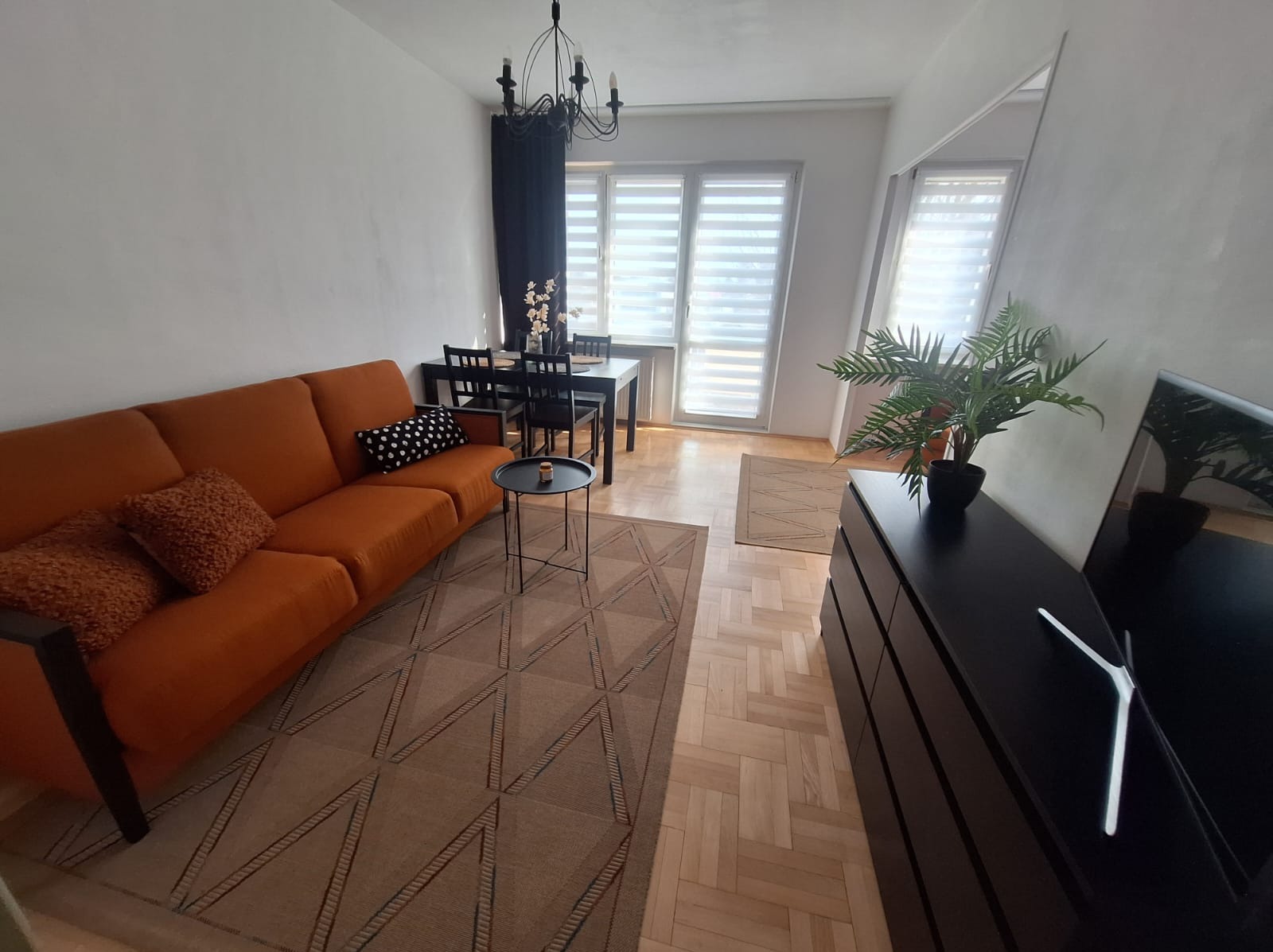 Salon w Kraczkowej: pomarańczowa sofa, wzorzysty dywan, czarne meble jadalne pod żyrandolem. Roślina doniczkowa na komodzie i parkiet dodają przytulności.