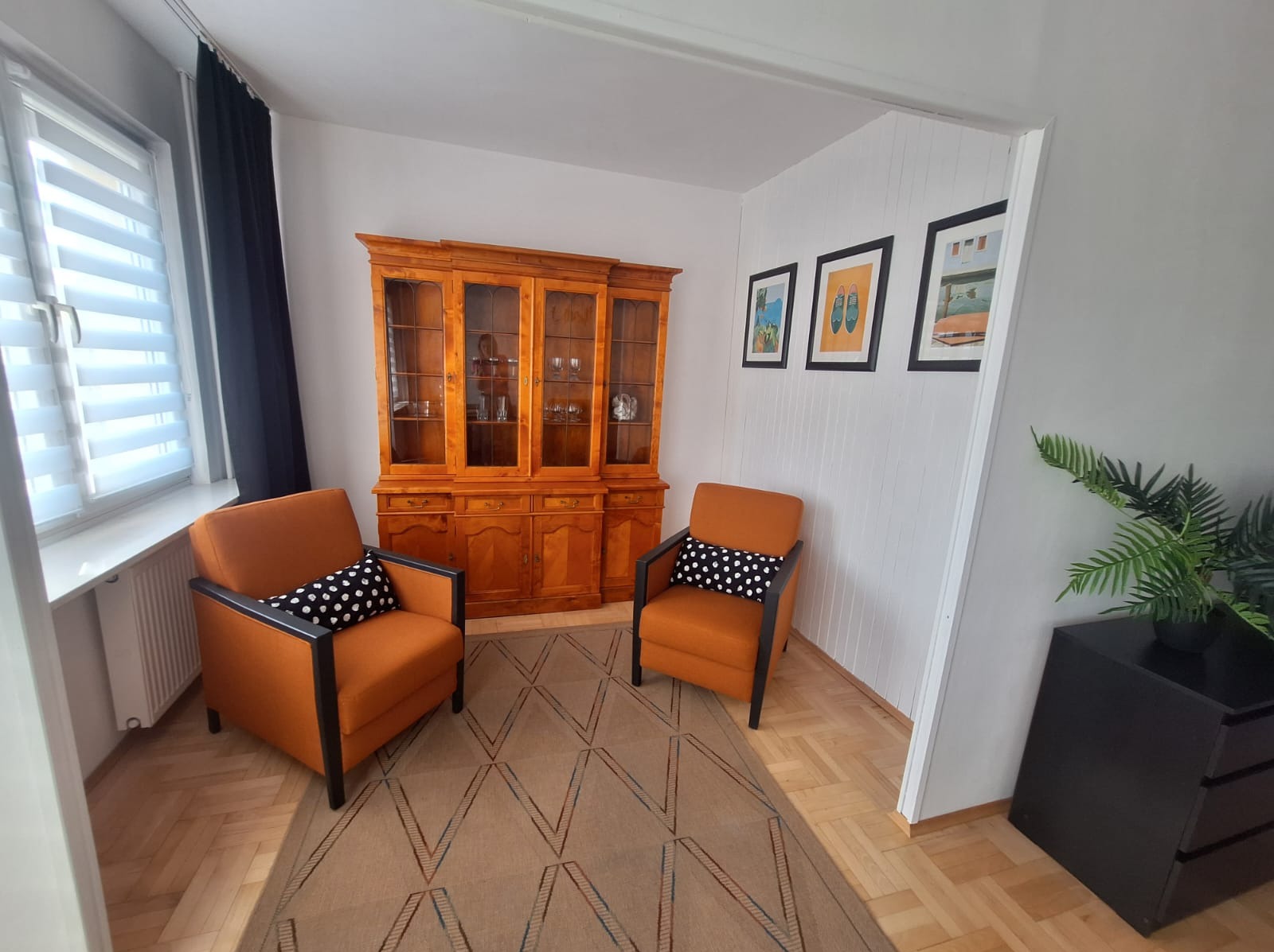 Dwa pomarańczowe fotele z czarnymi podłokietnikami i poduszkami w kropki, ustawione naprzeciwko siebie na wzorzystym dywanie, w tle drewniany kredens i obrazy.