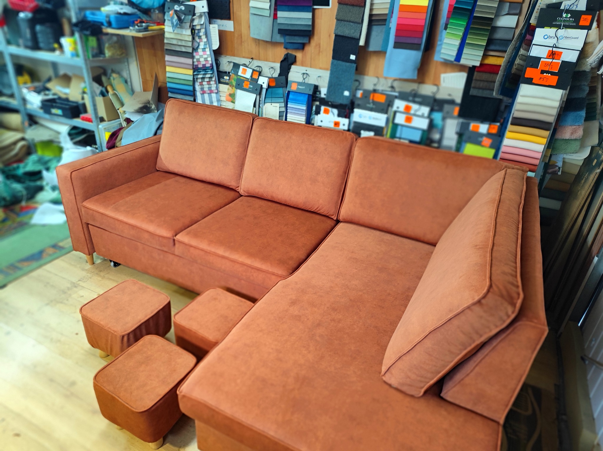 Narożnik z brązową tapicerką i pufami na tle próbek tkanin. Widoczne detale szycia i faktura materiału. Całość w ciepłej tonacji.