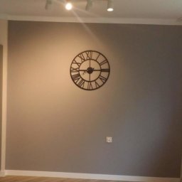 TopKolor - Ściana pomalowana na szaro z dekoracyjnym zegarem w stylu vintage, listwa przypodłogowa biała, oświetlenie punktowe na suficie, widoczny fragment szafki.